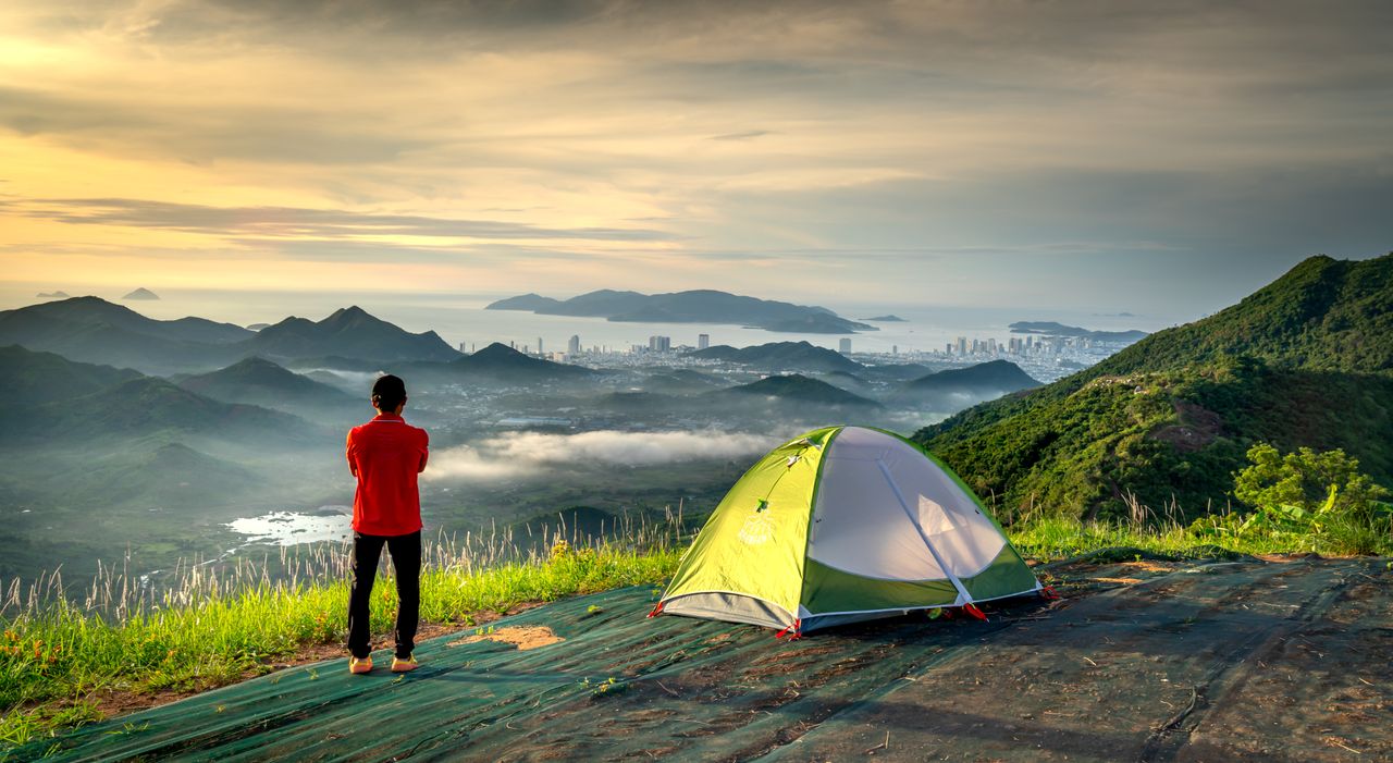 Home Adventurer stands by tent, embracing scenic mountain and city view at sunrise.